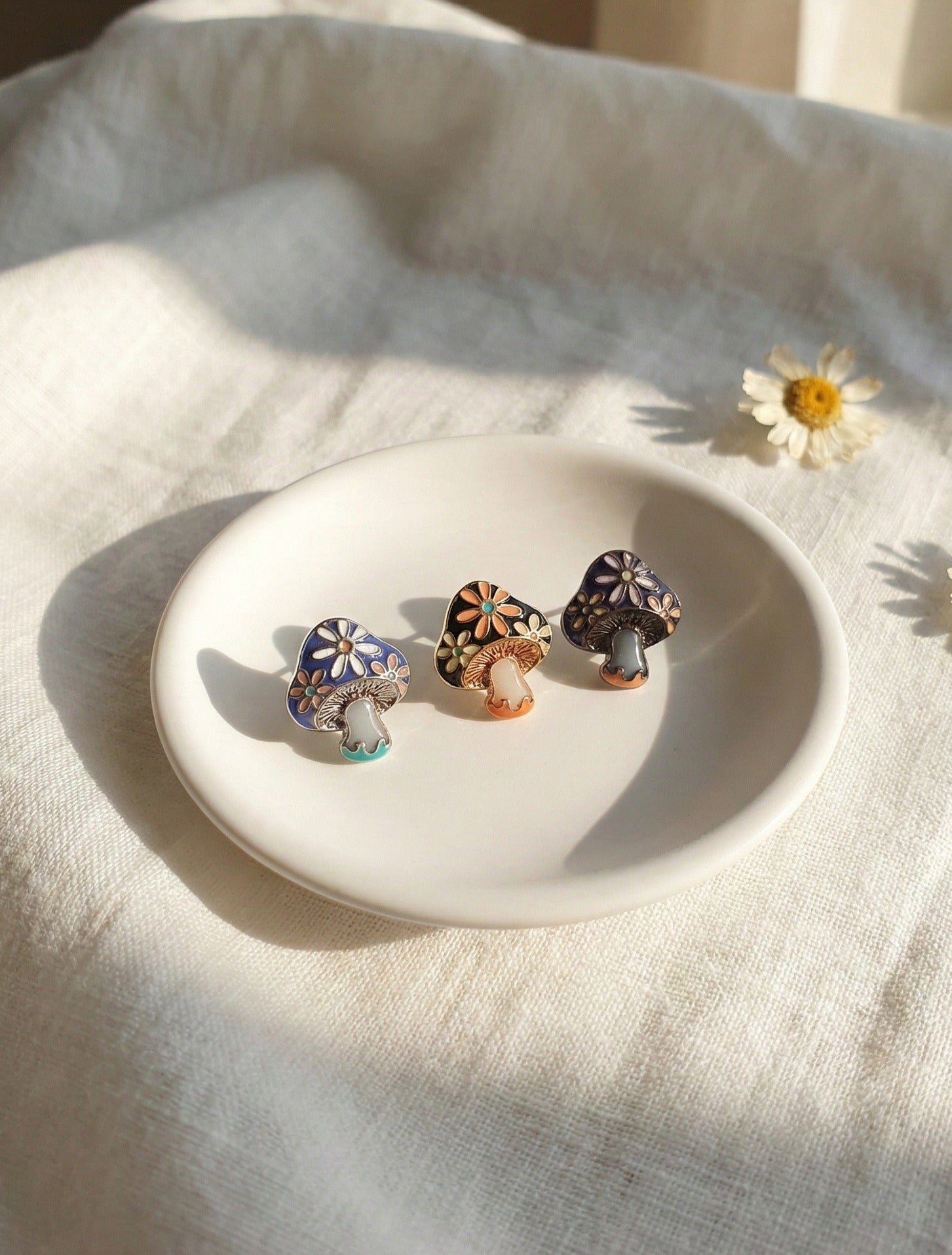 Three handmade enamel mushroom stud earrings with floral patterns on white ceramic plate with daisy flower