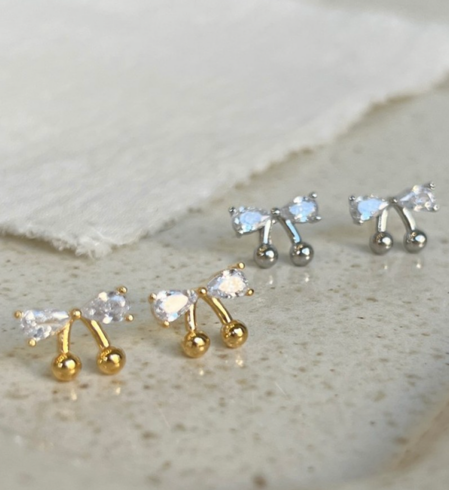 Gold and silver CZ bow cherry stud earrings displayed together on a stone surface, showing both colorways side by side