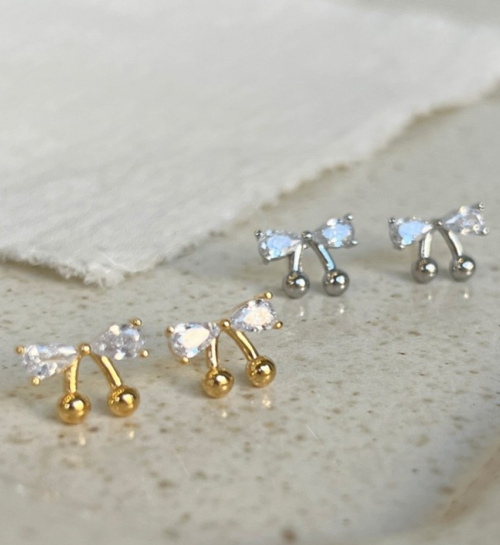 Gold and silver CZ bow cherry stud earrings displayed together on a stone surface, showing both colorways side by side