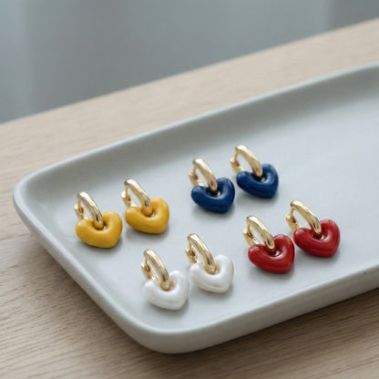 Four pairs of puffy heart huggie hoop earrings in red, navy, yellow, and pearl white colors displayed on a white ceramic tray