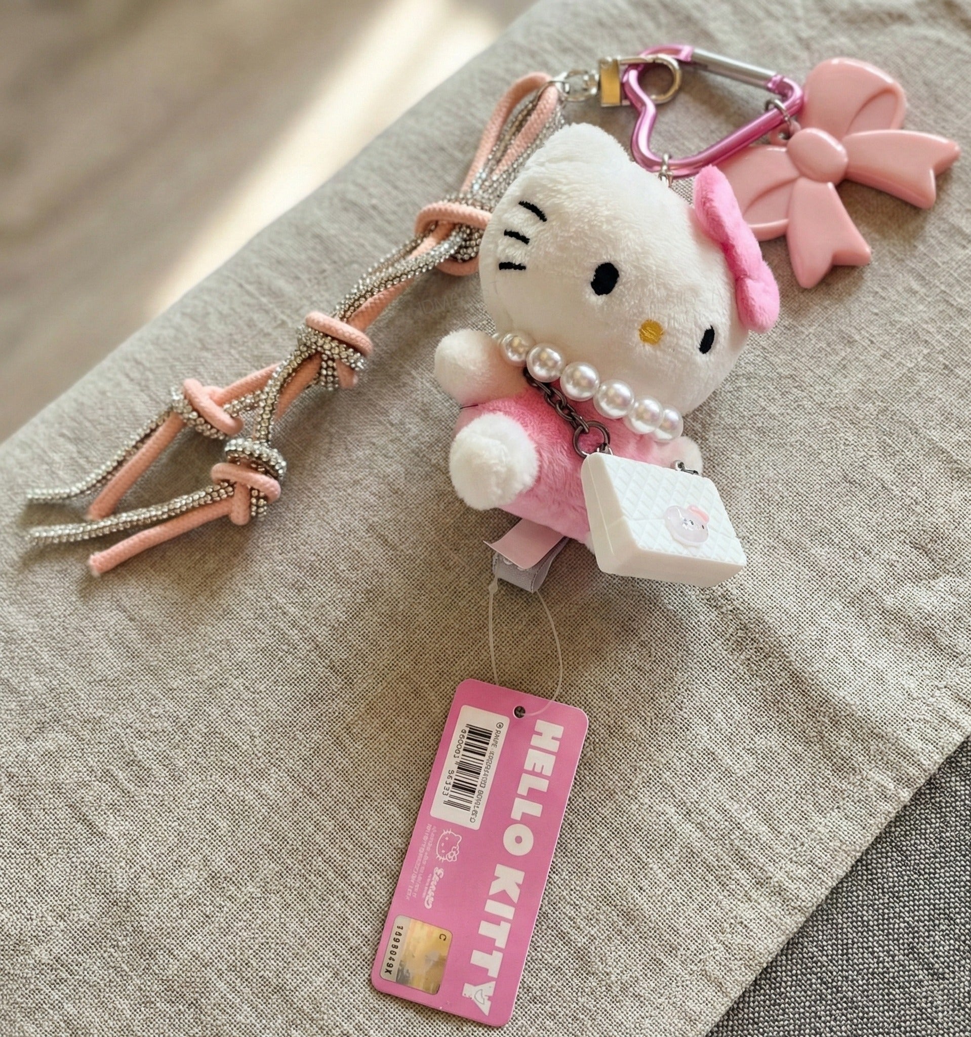 Authentic Sanrio Hello Kitty plush keychain with pearl necklace, quilted mini bag charm, crystal rhinestone rope strap, pink bow, and heart carabiner — official tag shown