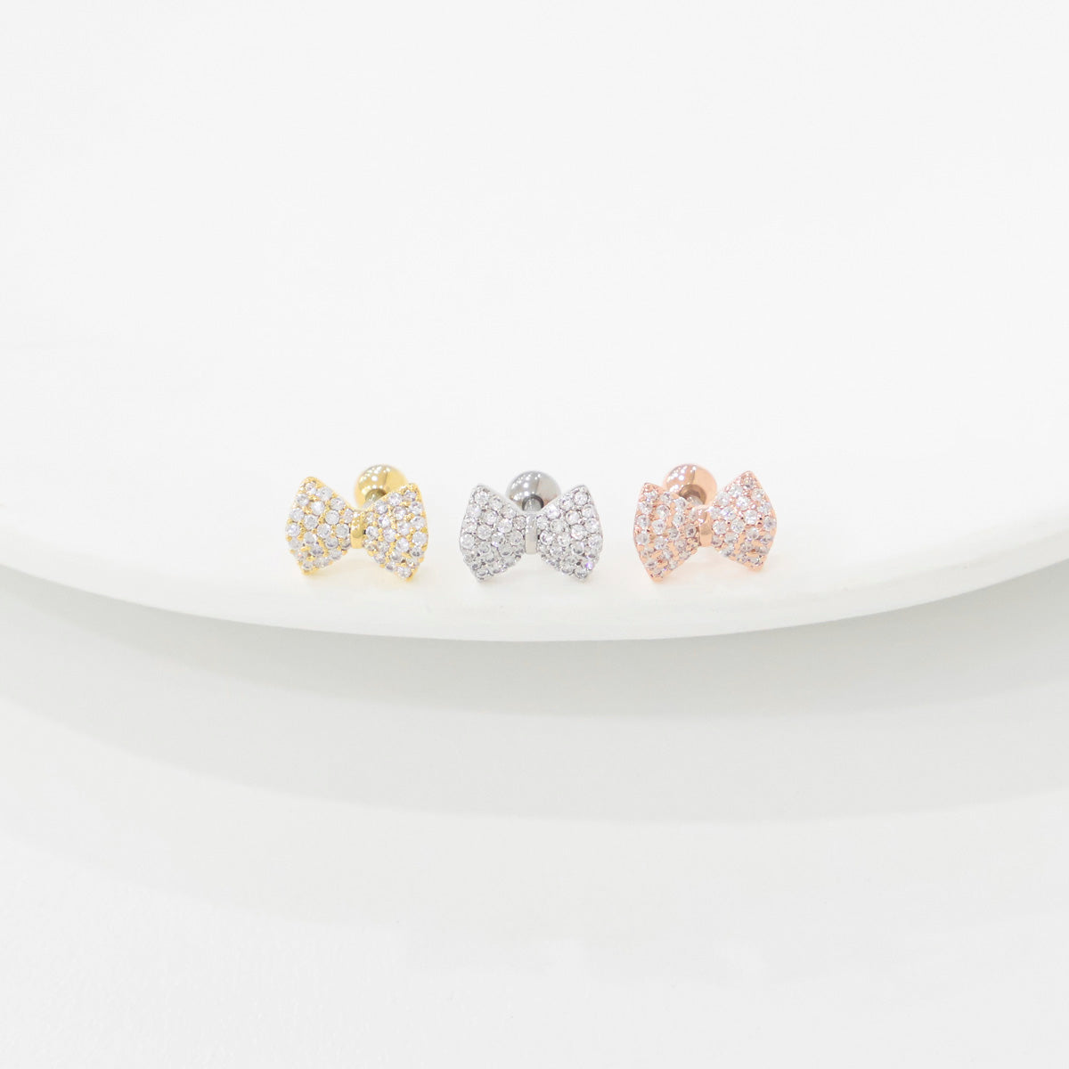 Three bow-shaped barbell earrings with cubic zirconia, shown in gold, silver, and rose gold, displayed on a white ceramic dish
