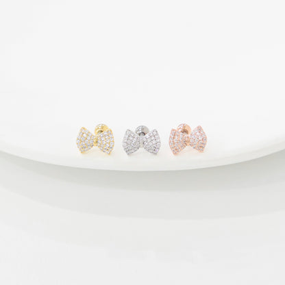 Three bow-shaped barbell earrings with cubic zirconia, shown in gold, silver, and rose gold, displayed on a white ceramic dish