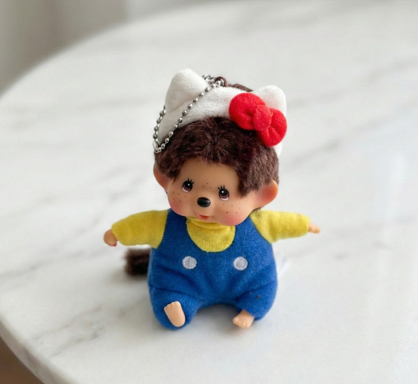 Monchhichi plush keychain wearing Hello Kitty red bow headband with blue overalls on white surface