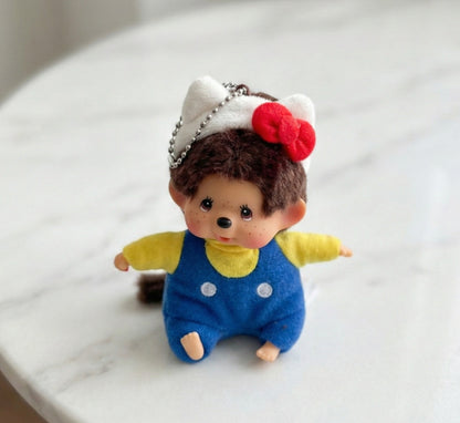 Monchhichi plush keychain wearing Hello Kitty red bow headband with blue overalls on white surface