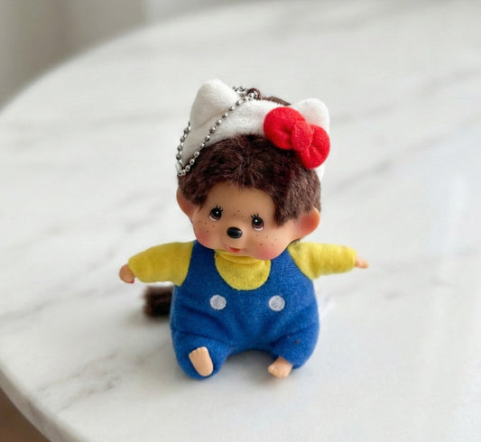 Monchhichi plush keychain wearing Hello Kitty red bow headband with blue overalls on white surface