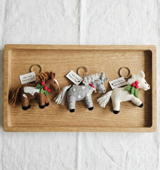 Three handmade wool felt pony keychains in brown, grey dot, and white colors displayed on wooden tray with '100% Wool Made in Nepal' tags