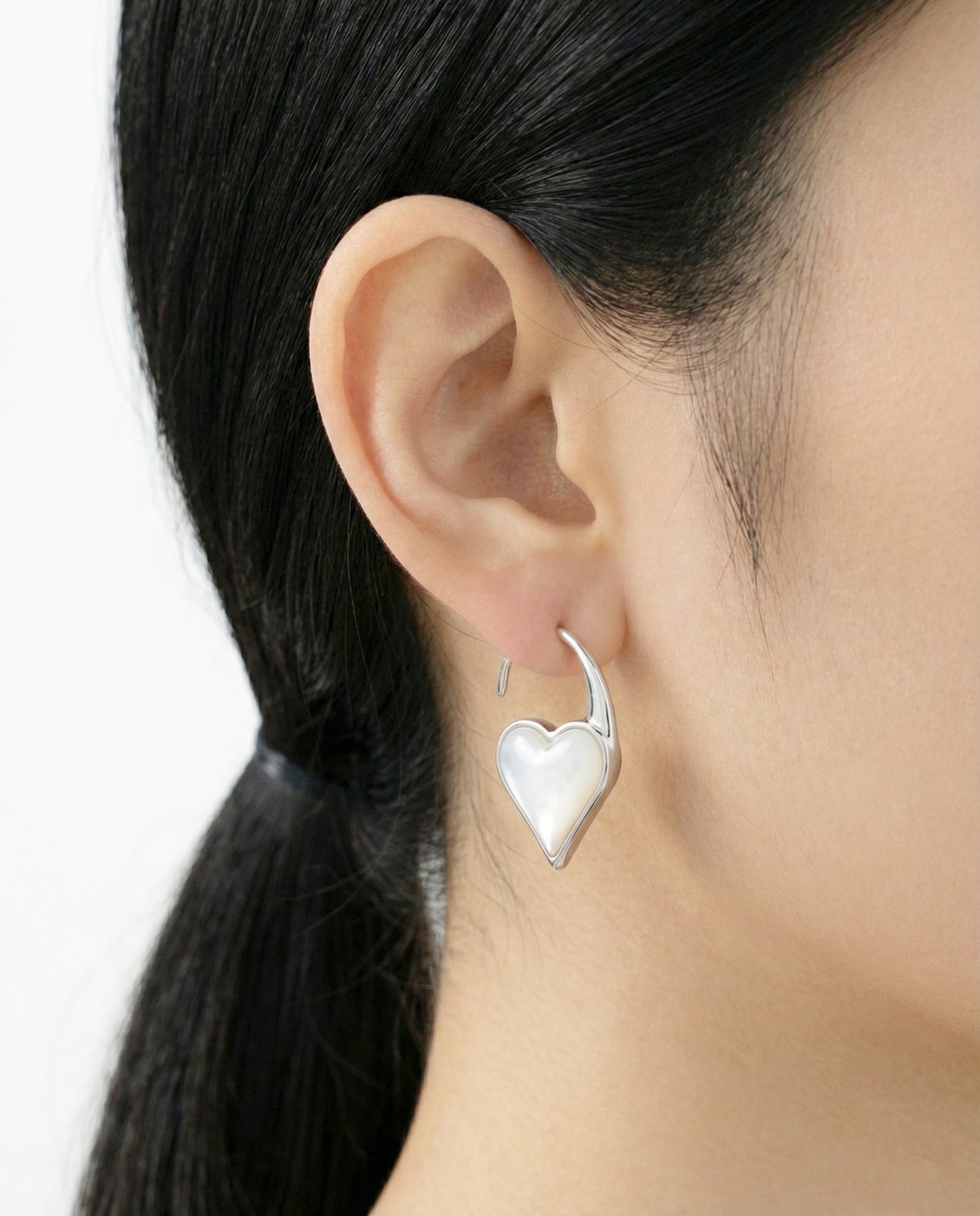 Close-up of a woman wearing a silver heart hook earring with a glossy mother-of-pearl inlay, styled with sleek black hair