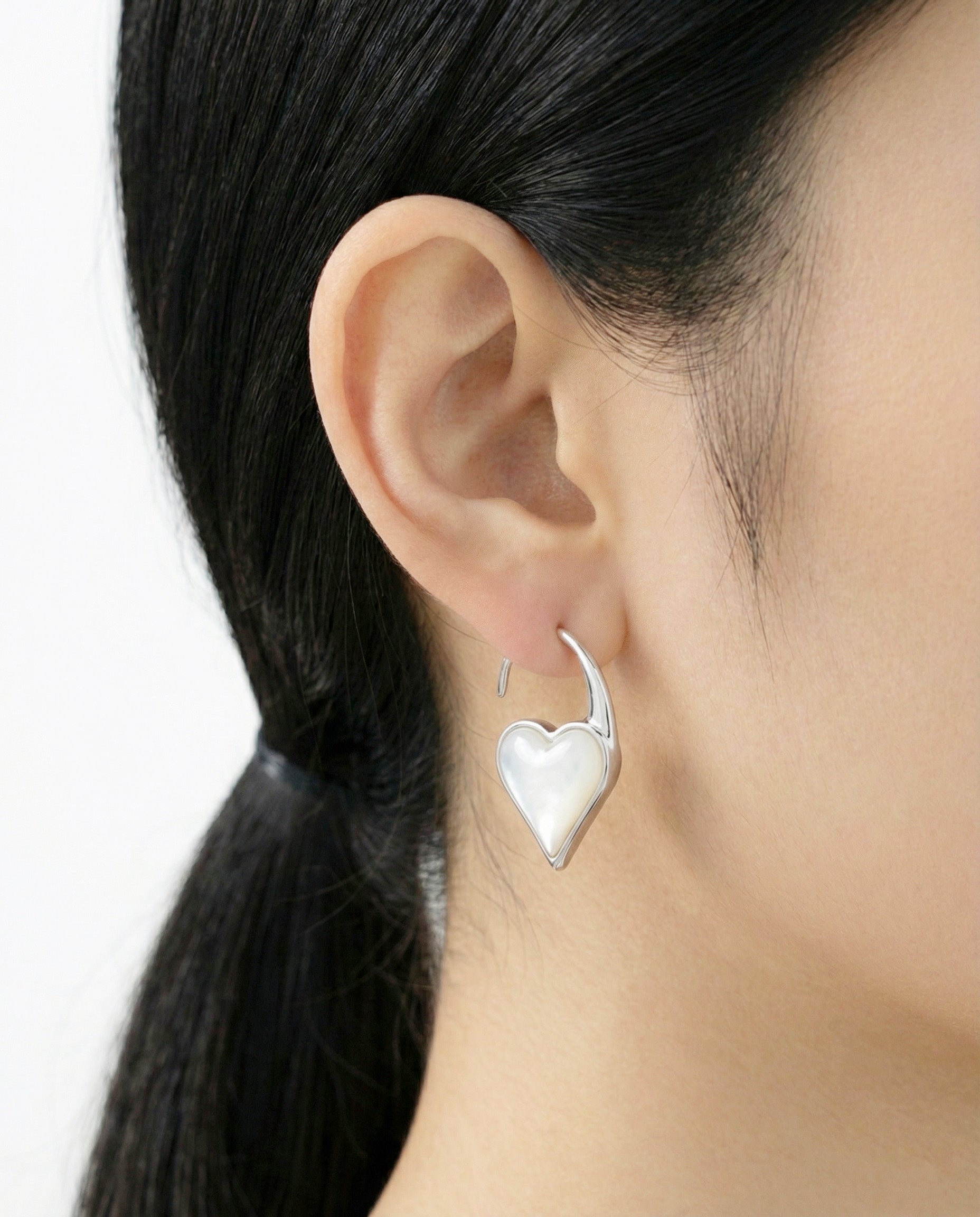 Close-up of a woman wearing a silver heart hook earring with a glossy mother-of-pearl inlay, styled with sleek black hair