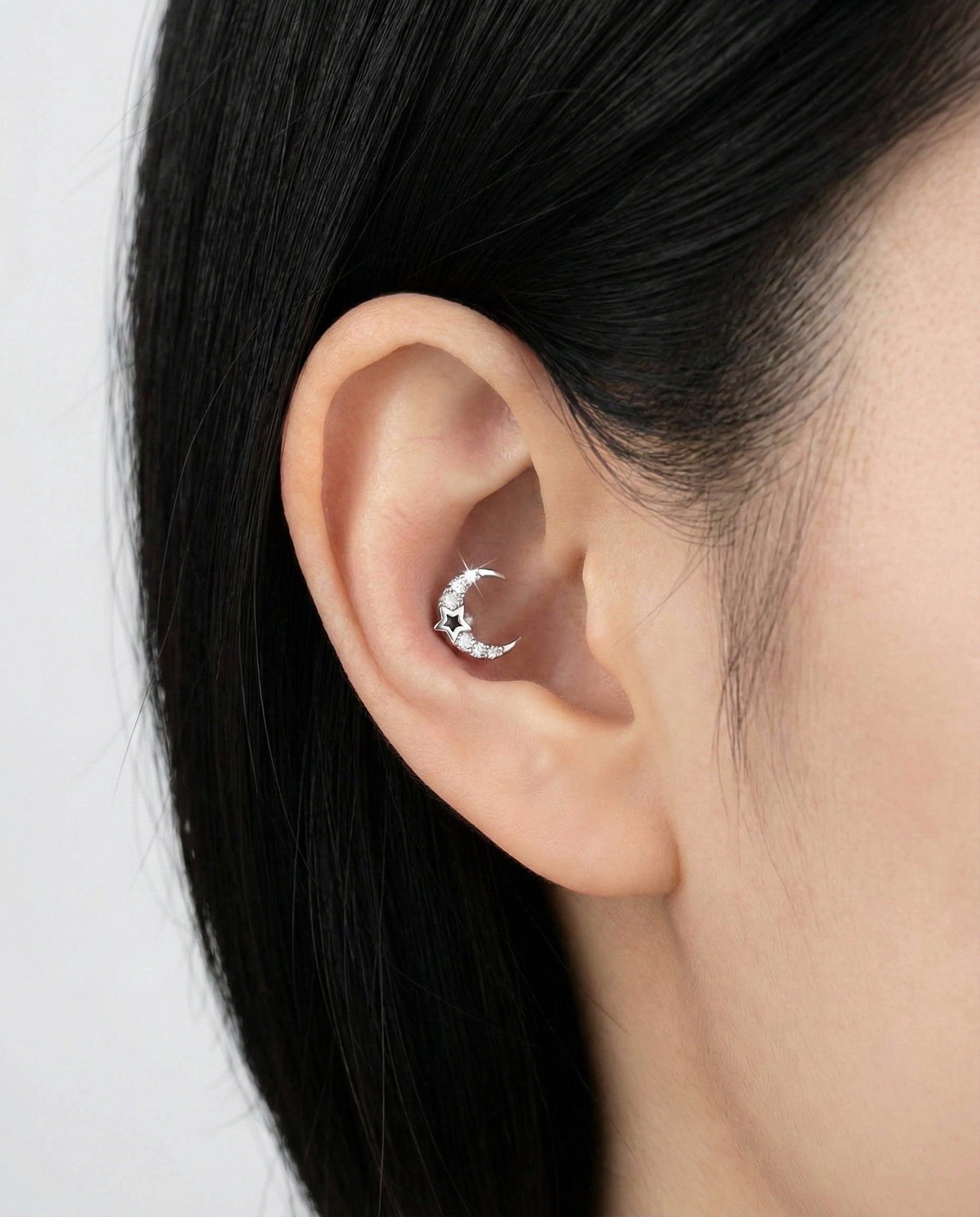 Close-up of a silver crescent moon cartilage earring with embedded clear crystals and a black star cutout, worn on the inner ear.