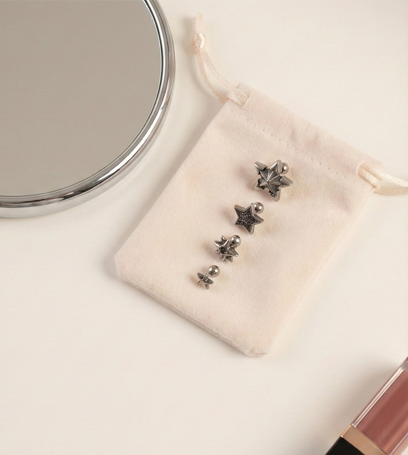 Four vintage antique star barbell piercings displayed on a cream velvet pouch next to a round mirror and pink lip gloss