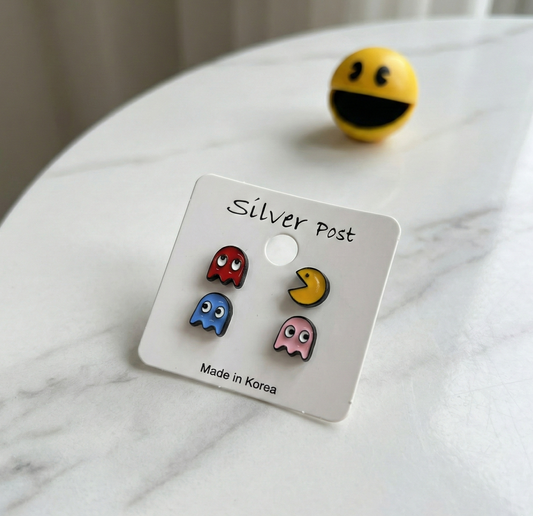 Pac-Man and Ghost enamel stud earring set on display card labeled 'Silver Post / Made in Korea', with a yellow Pac-Man toy ball in background on marble surface