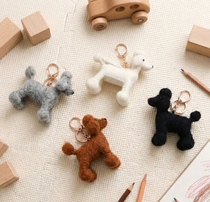 Four handmade needle felted wool poodle keychains in white, brown, grey, and black colors displayed on cream fabric with wooden toys