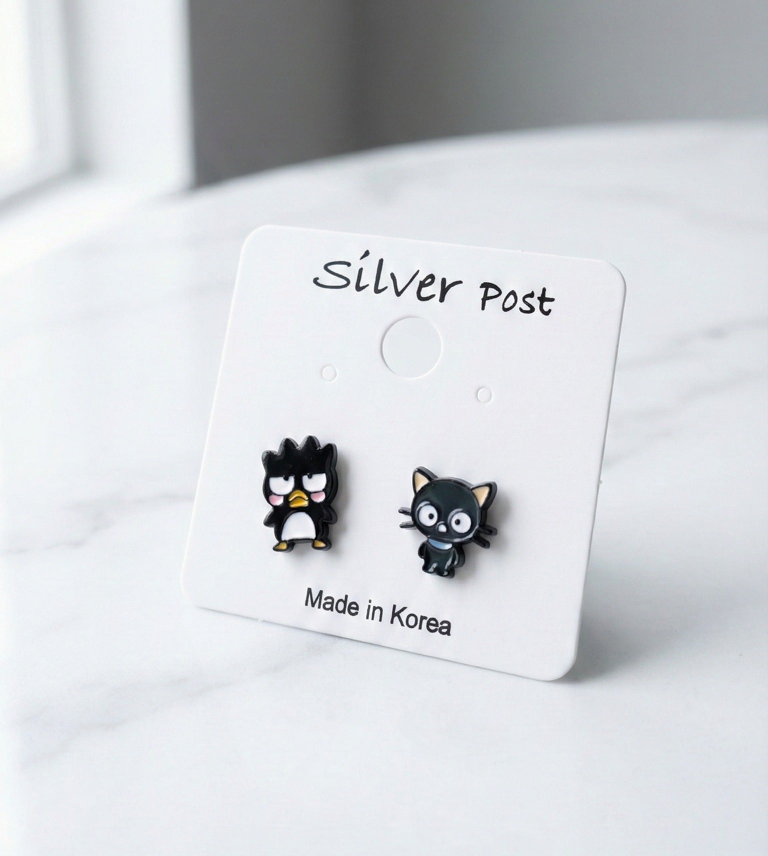Close up of Badtz-Maru penguin and Chococat black cat stud earrings on white display card with silver post label, handmade in Korea
