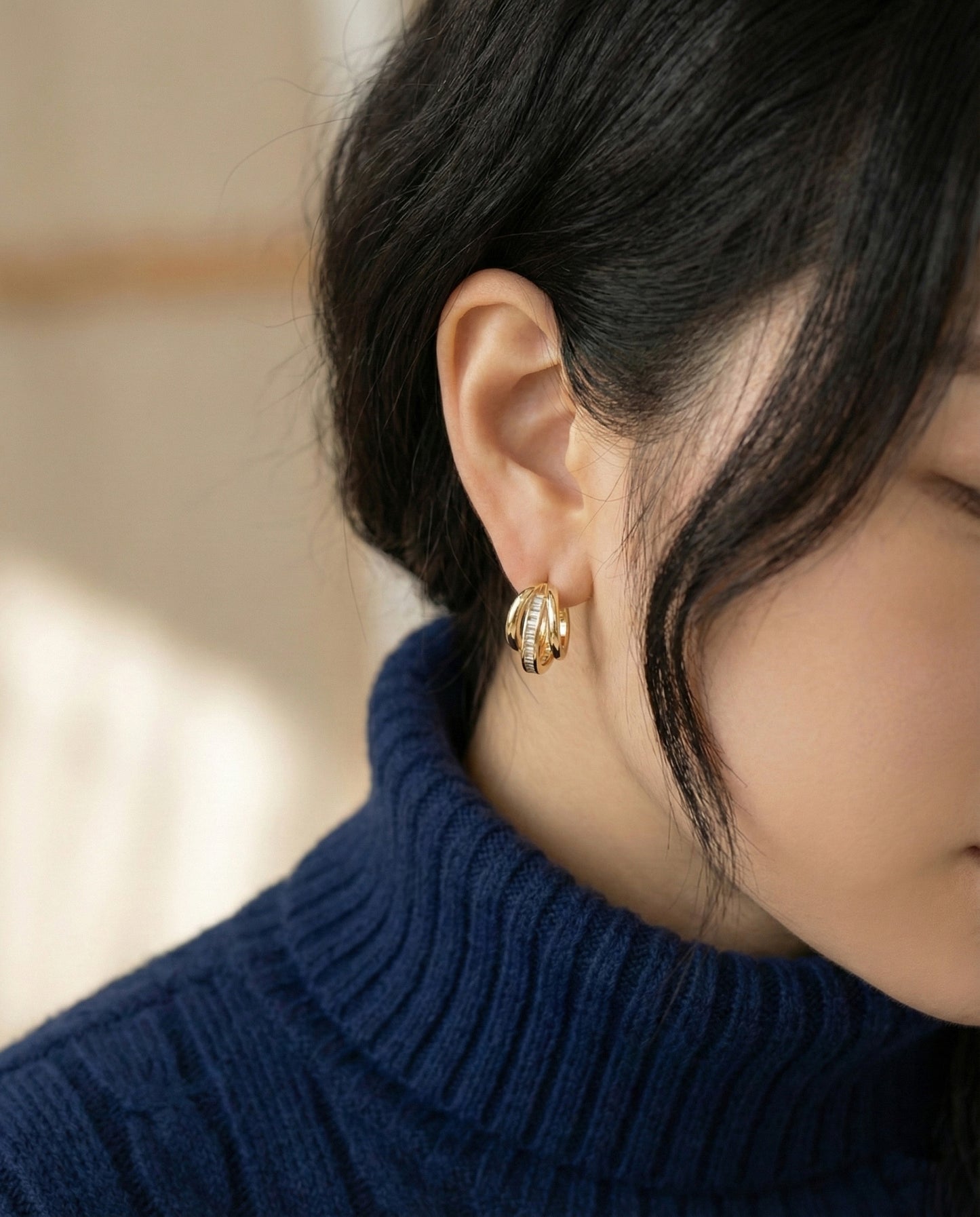 Woman wearing gold triple row baguette huggie hoop earrings with navy turtleneck sweate