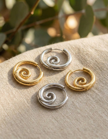 Gold and silver spiral hoop earrings displayed on beige linen fabric with eucalyptus leaves