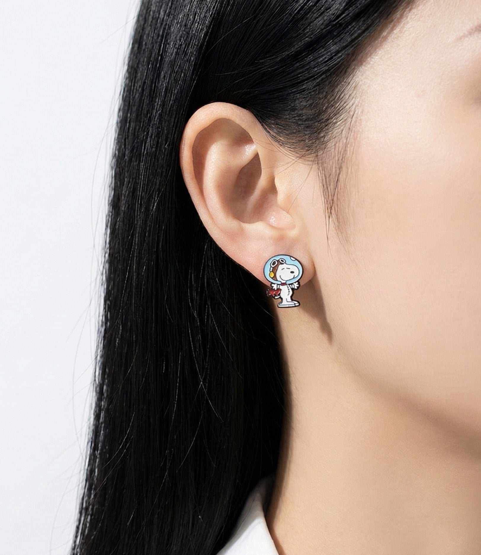 Close-up of a woman wearing a silver post earring featuring Snoopy in a blue astronaut helmet, facing forward.