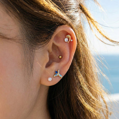 Close-up of ear wearing dolphin and whale tail barbell stud earrings with crystal and blue gem accents