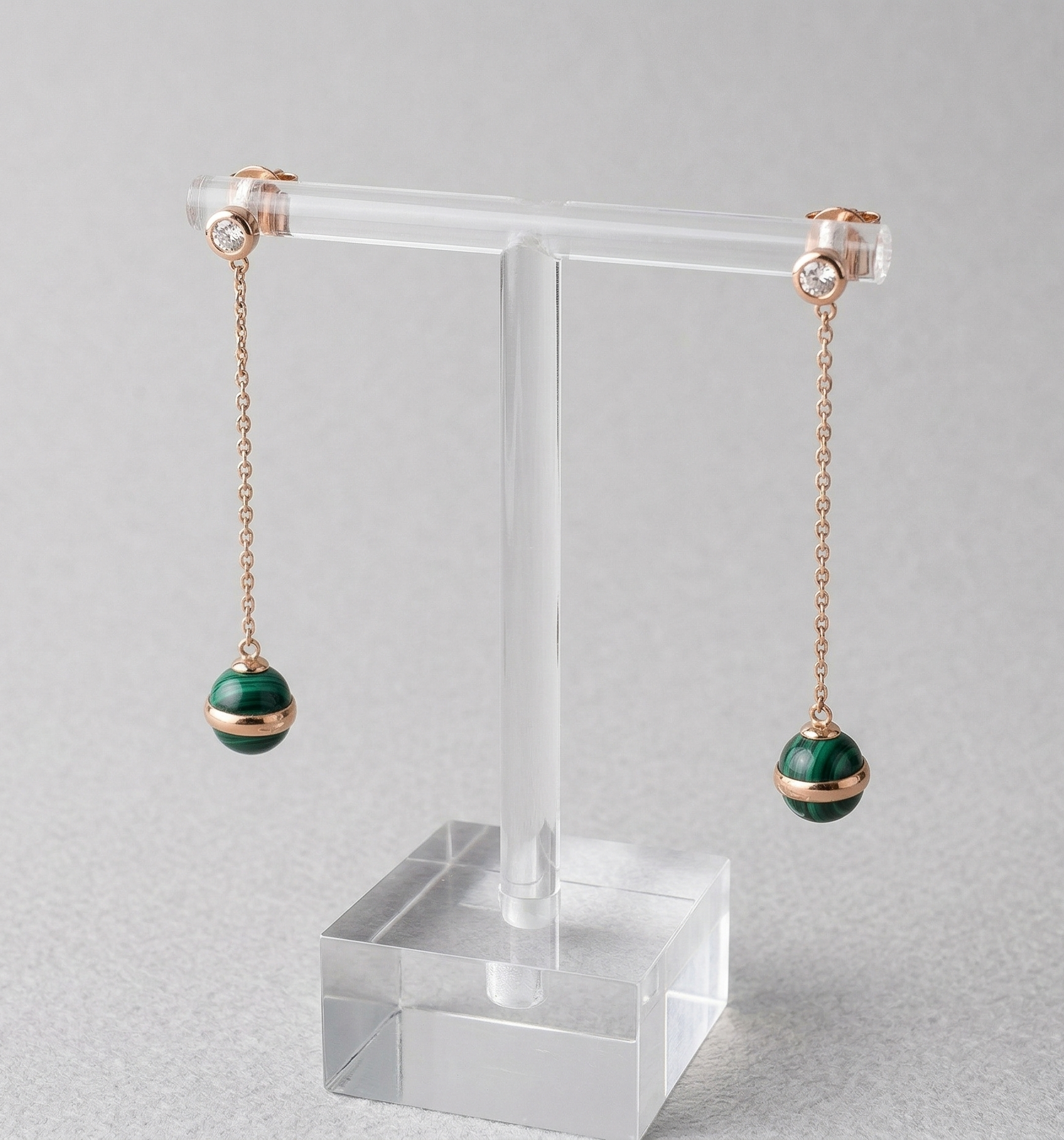 Natural malachite ball drop earrings displayed on acrylic T-bar stand, showing rose gold chain, CZ bezel stud top, and 8mm green malachite sphere with saturn-ring band detail