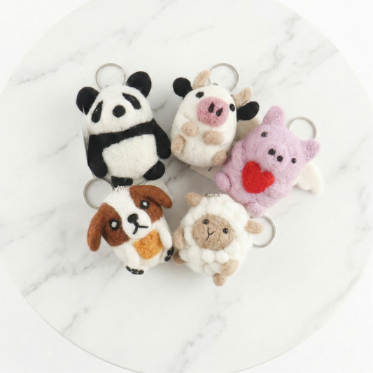 Five handmade Nepal wool felt animal keychains including panda, cow, pig, dog, and sheep on white marble background"