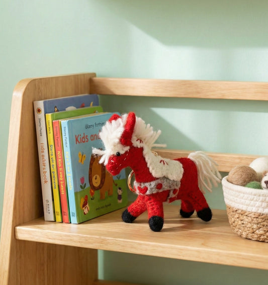Handmade red wool felt horse keychain displayed on wooden bookshelf with children's books, nursery decor style