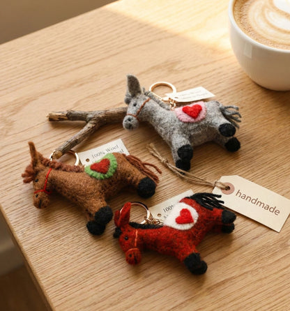 Three handmade wool felt horse keychains in brown, red, and grey displayed on wooden tray with coffee