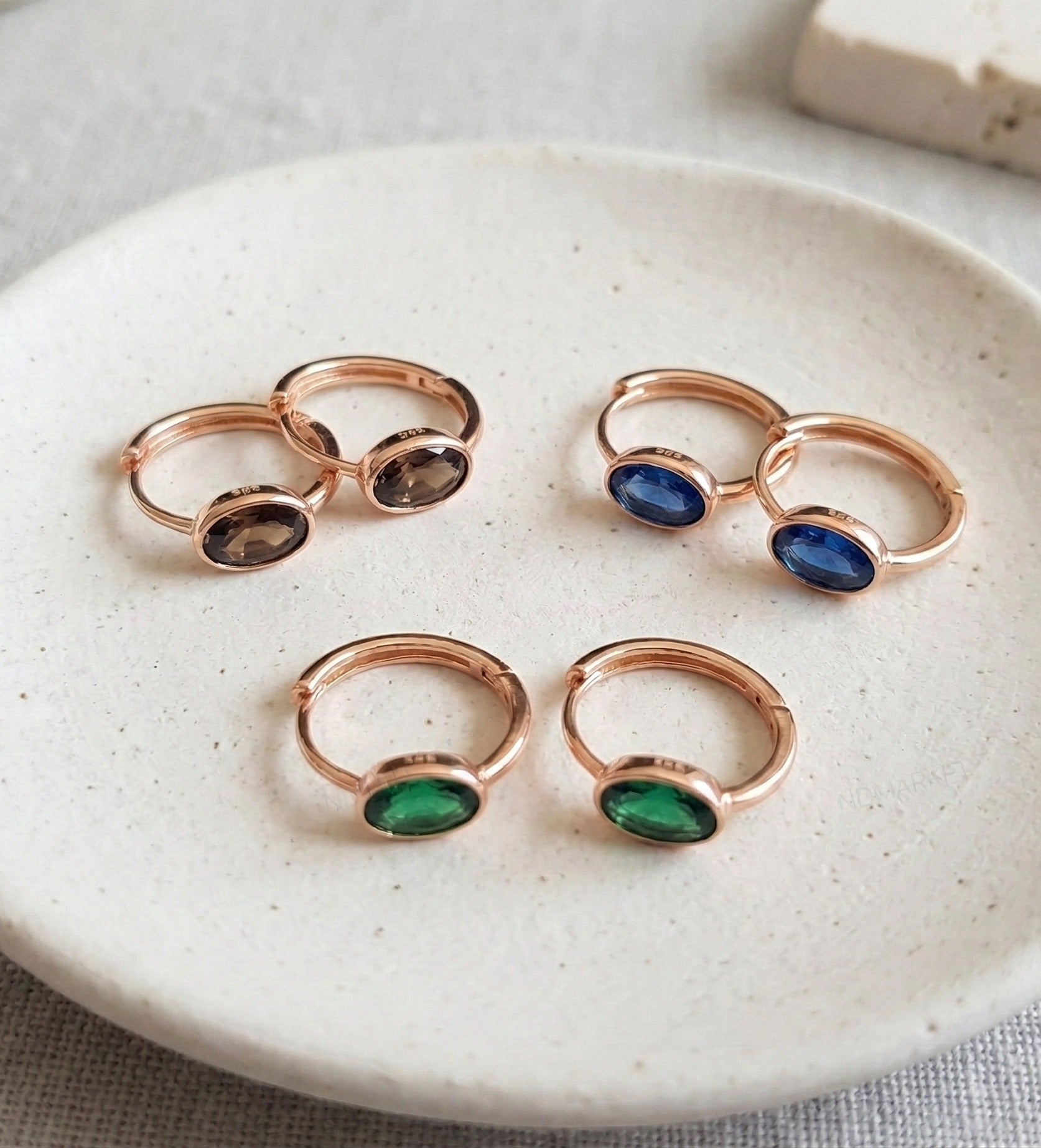 :K-style jewelry flat lay featuring Oval Bezel CZ Huggie Earrings in emerald, sapphire and smoky rose gold, hypoallergenic 925 silver all three colorways.