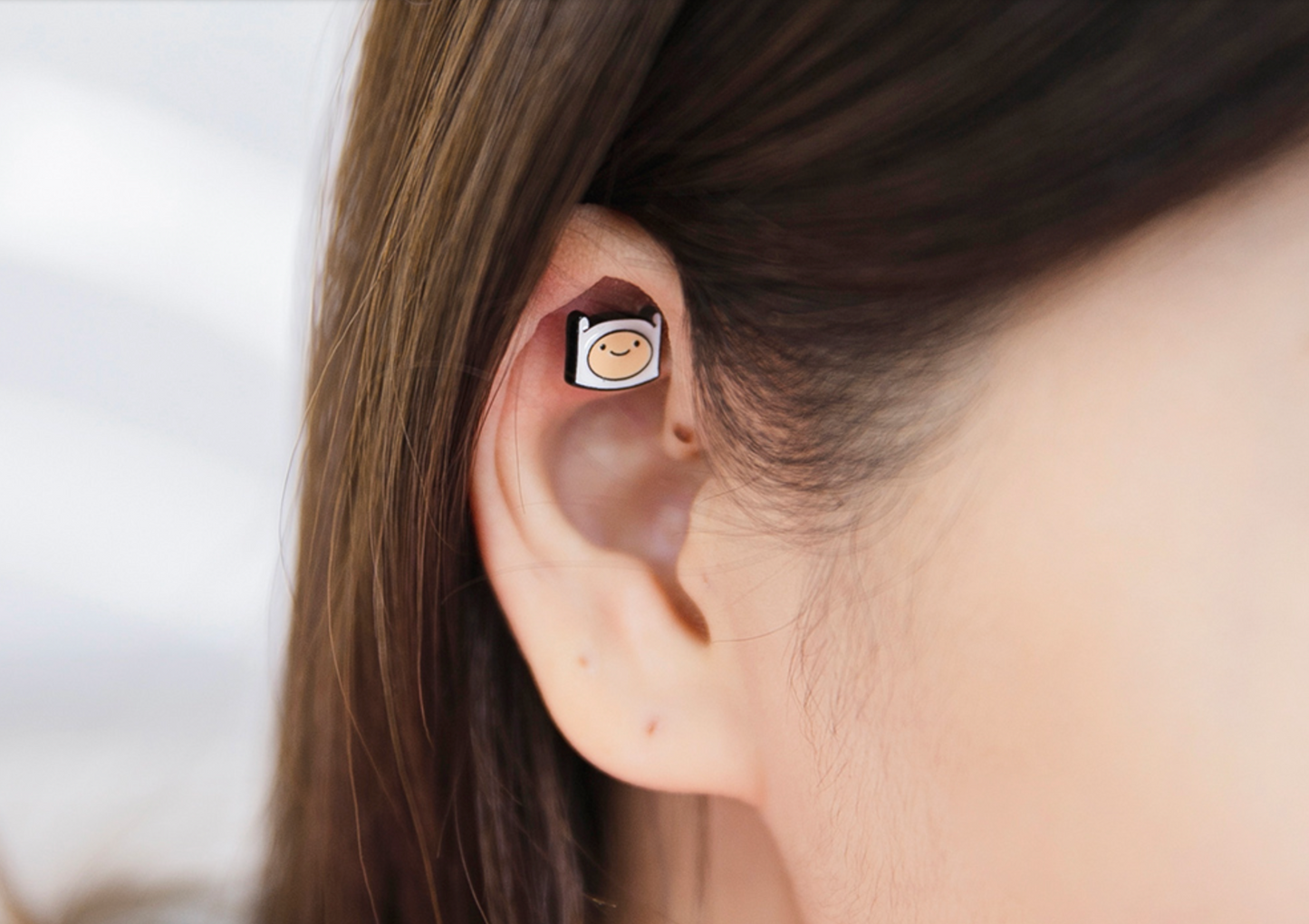 Close-up of a person wearing an Adventure Time Finn enamel barbell earring on the inner ear cartilage, smiling face design.