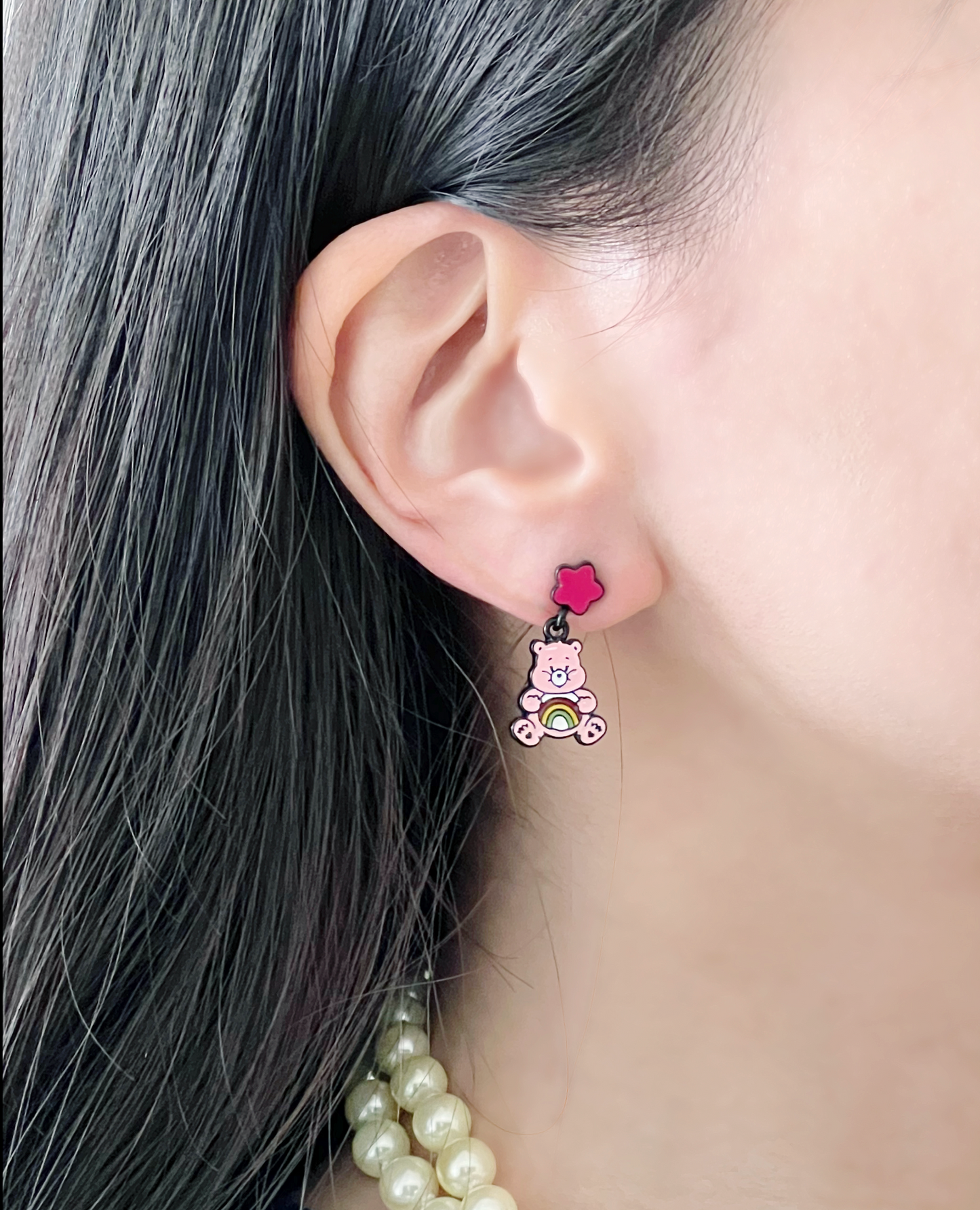 Close-up of a pink enamel bear earring with a rainbow belly and a flower-shaped pink stud, worn with a pearl necklace.