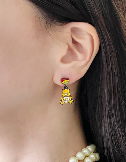 A yellow enamel bear earring with a sun belly and a rainbow-striped heart stud, worn with hair tucked behind the ear.