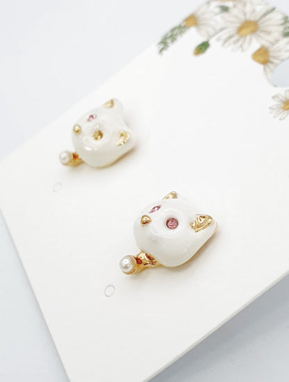 White enamel cat-shaped stud earrings with pink crystal eyes, gold ear tips, and a tiny pearl drop on the bottom.