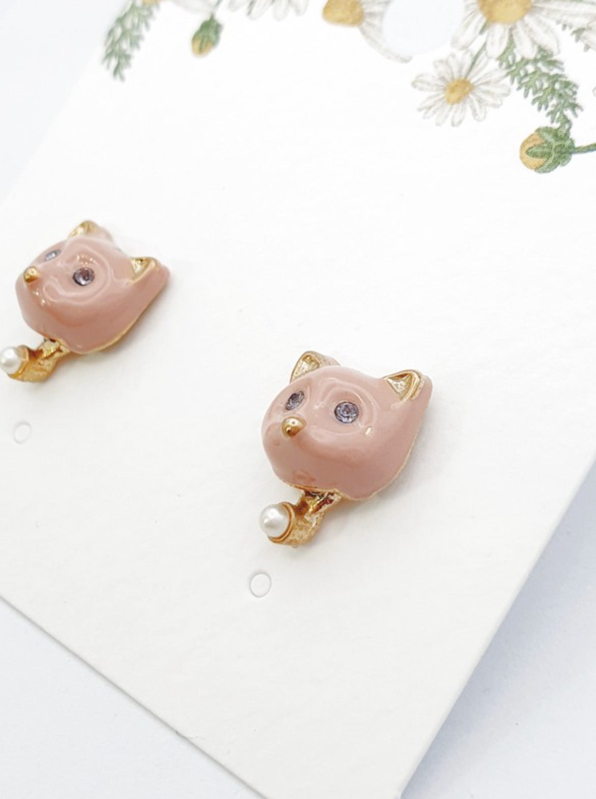 Soft pink enamel cat-shaped stud earrings with lavender crystal eyes, gold ear tips, and a tiny pearl drop detail.