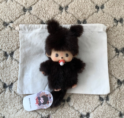 Brown fuzzy Monchhichi-style plush doll with pacifier, lying flat on a white drawstring pouch atop a textured cream rug