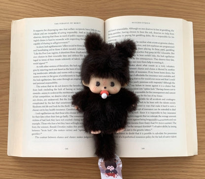 Furry black doll with red pacifier laid on top of an open English book, styled as a cozy desk or study scene.
