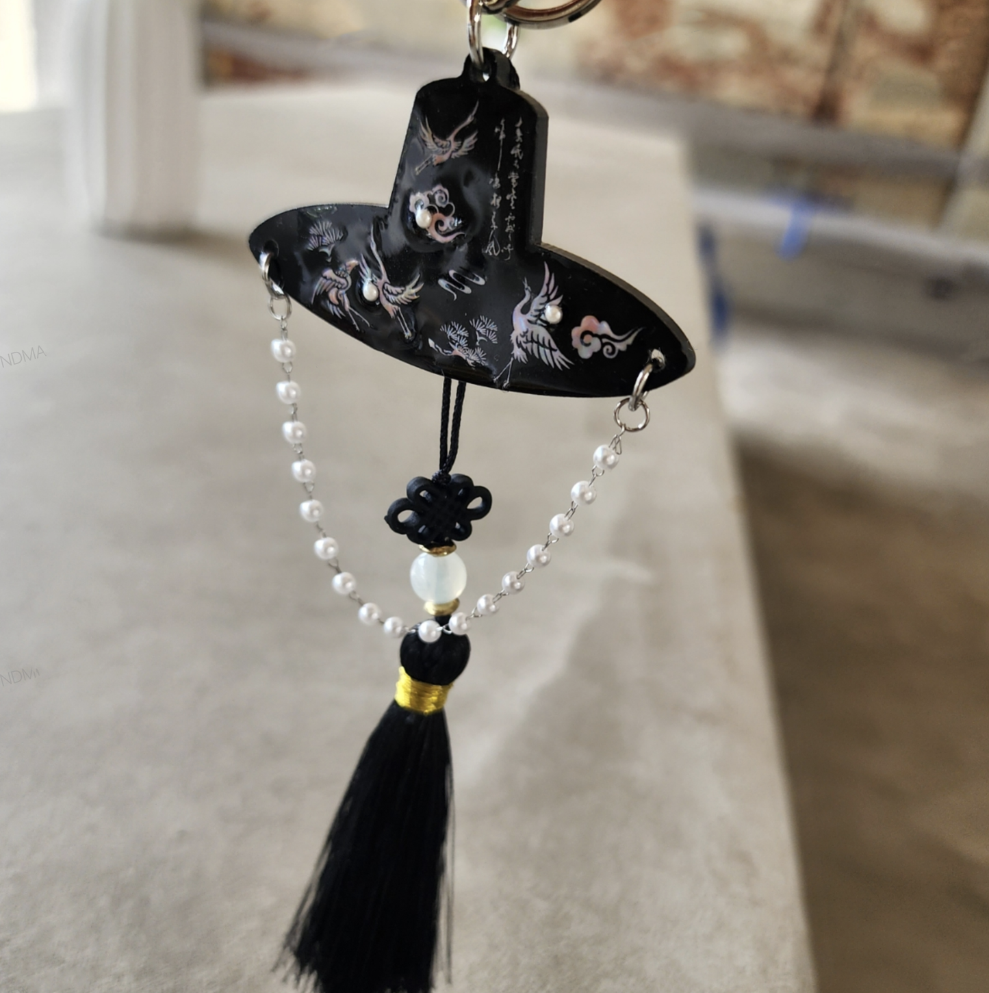 Single black tassel keychain with Korean traditional hat motif and pearl strands, held by hand in front of a hanok house.