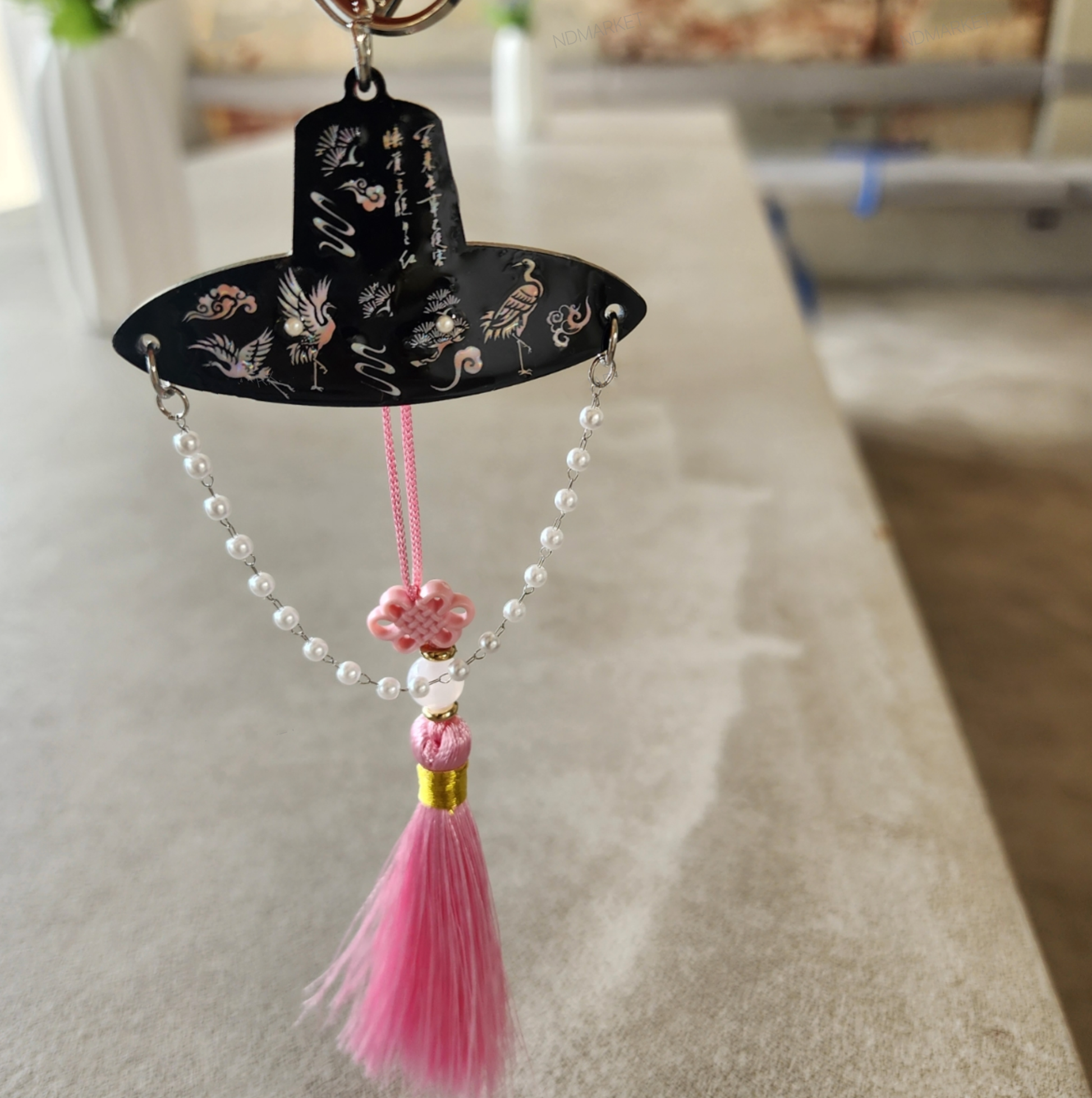 Pink tassel keychain styled as a Korean traditional hat with floral knots and pearl strands, shown in front of a blurred hanok backdrop.