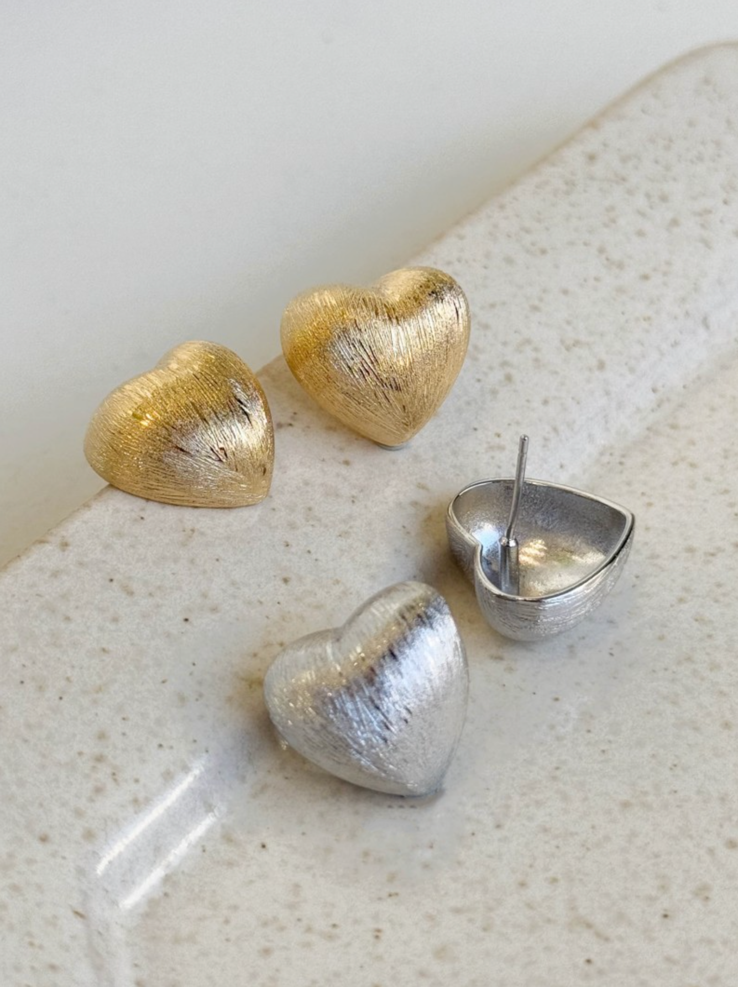 gold and silver brushed-metal heart stud earrings displayed on a speckled stone tray, including one open back showing the post.
