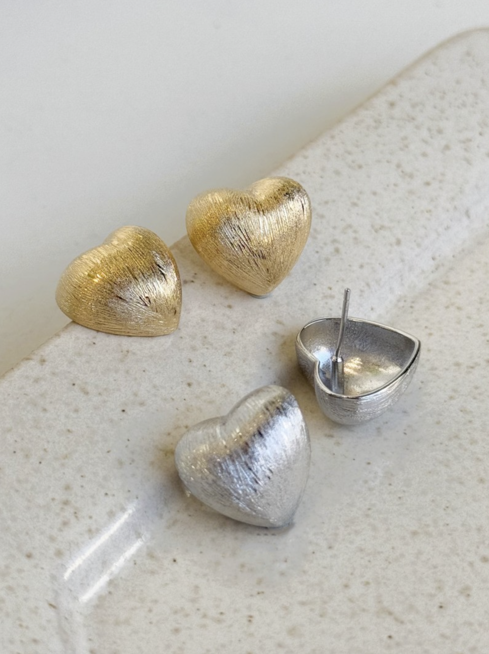 gold and silver brushed-metal heart stud earrings displayed on a speckled stone tray, including one open back showing the post.
