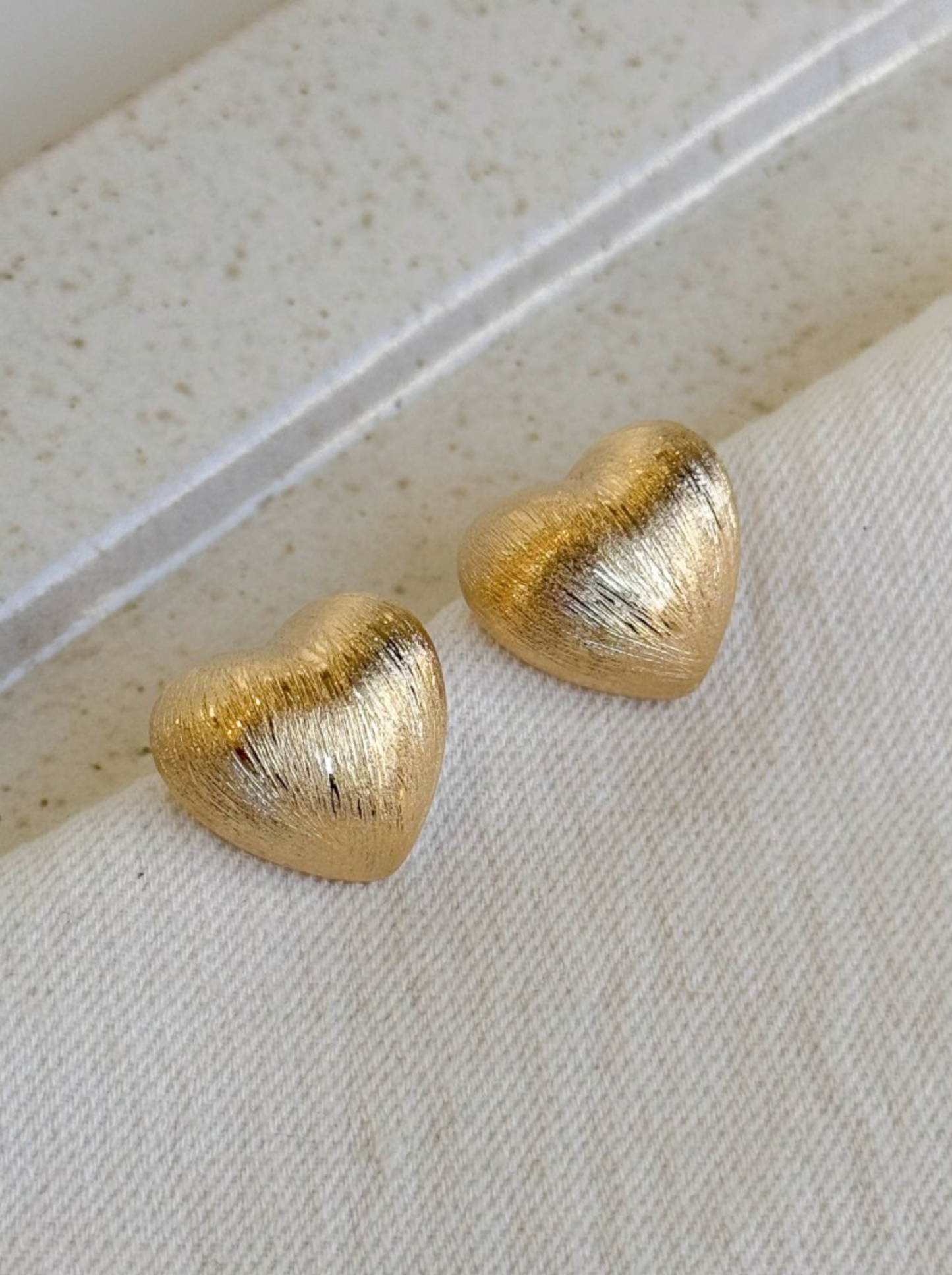 gold brushed-finish heart stud earrings placed on a folded fabric edge under soft natural light.