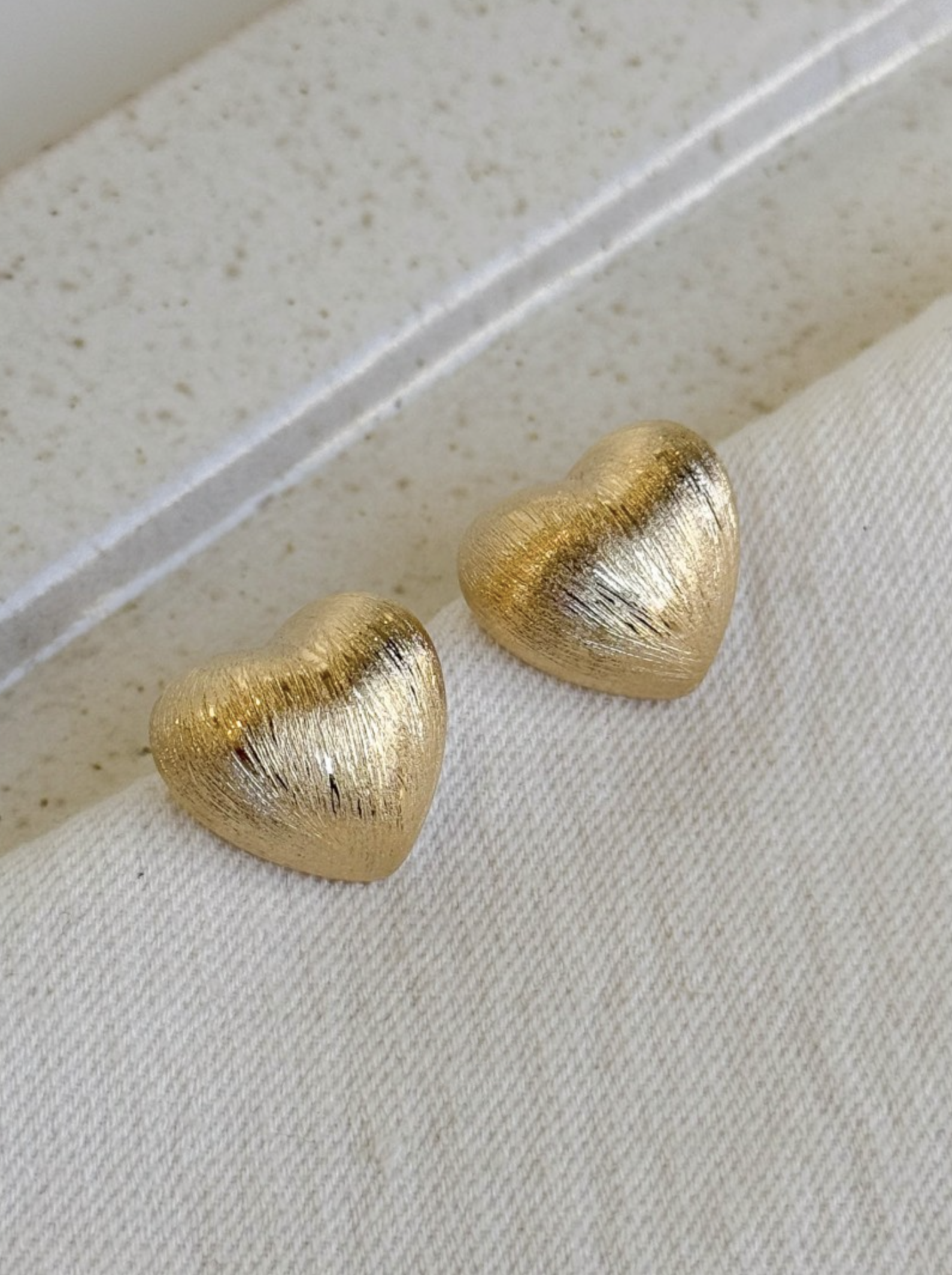 gold brushed-finish heart stud earrings placed on a folded fabric edge under soft natural light.