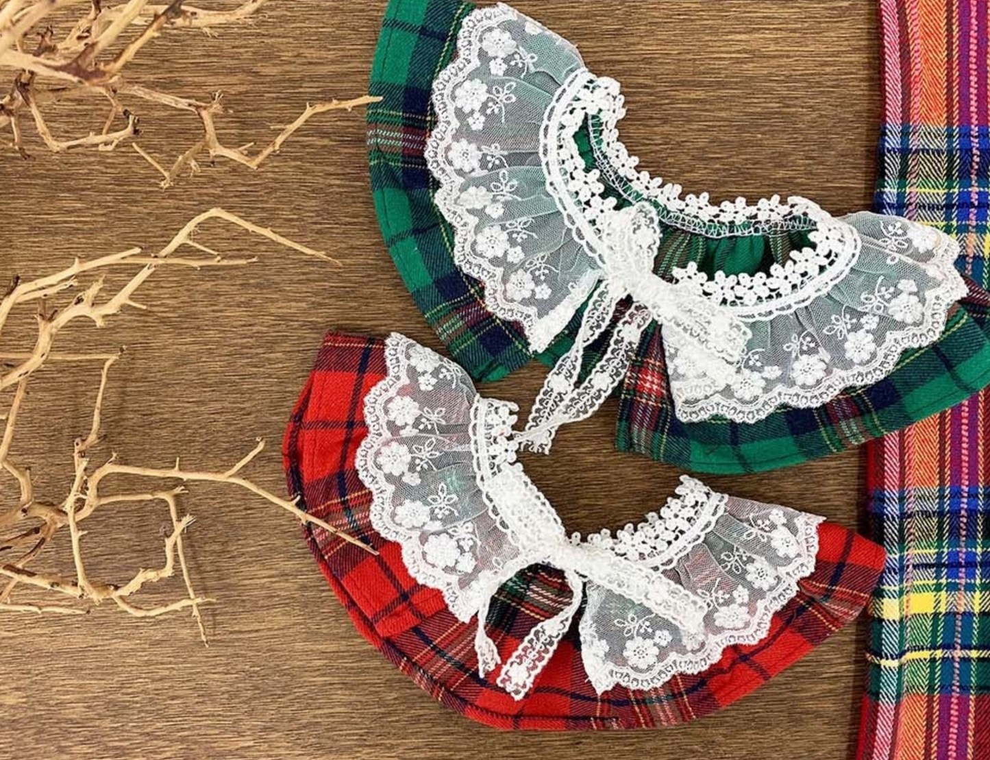 Two tartan dog capes — one red and one green — both with frilly white lace and bow ties. Placed flat side by side to show color variations.