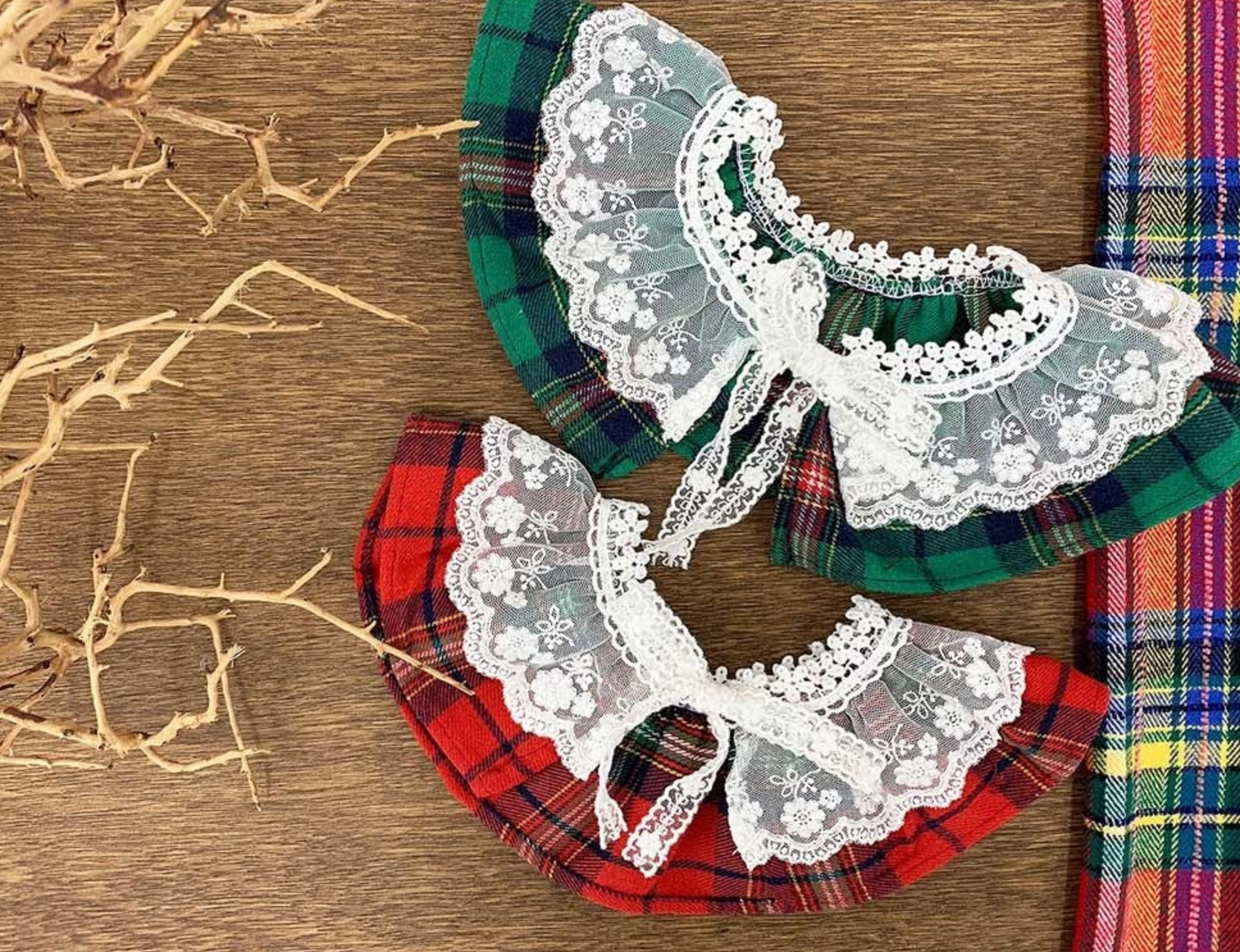 Two tartan dog capes — one red and one green — both with frilly white lace and bow ties. Placed flat side by side to show color variations.