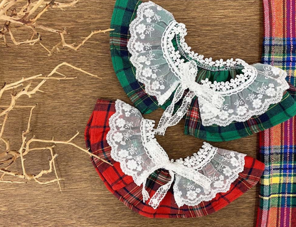 Two tartan dog capes — one red and one green — both with frilly white lace and bow ties. Placed flat side by side to show color variations.