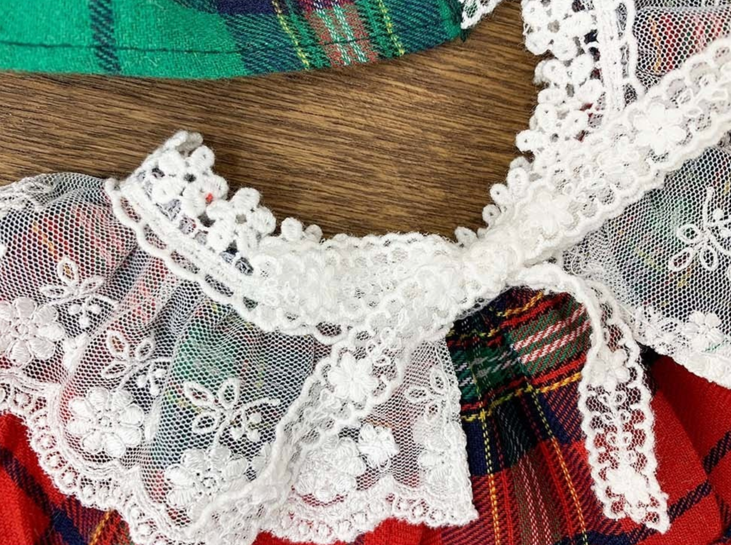 Close-up view of a red tartan dog cape featuring white floral lace trim and a delicate lace bow at the center. The texture and stitch detail are clearly visible.