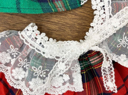 Close-up view of a red tartan dog cape featuring white floral lace trim and a delicate lace bow at the center. The texture and stitch detail are clearly visible.