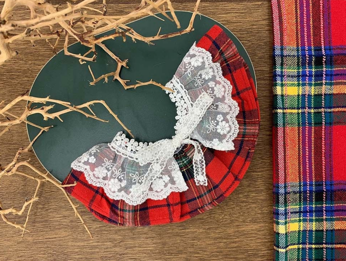 A red plaid dog cape with a frilly white lace collar, styled on a dark green plate with dry decorative branches. A matching red plaid fabric lies beside it.