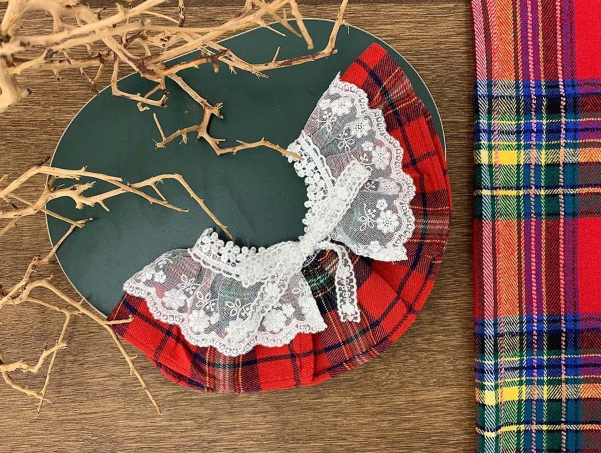 A red plaid dog cape with a frilly white lace collar, styled on a dark green plate with dry decorative branches. A matching red plaid fabric lies beside it.