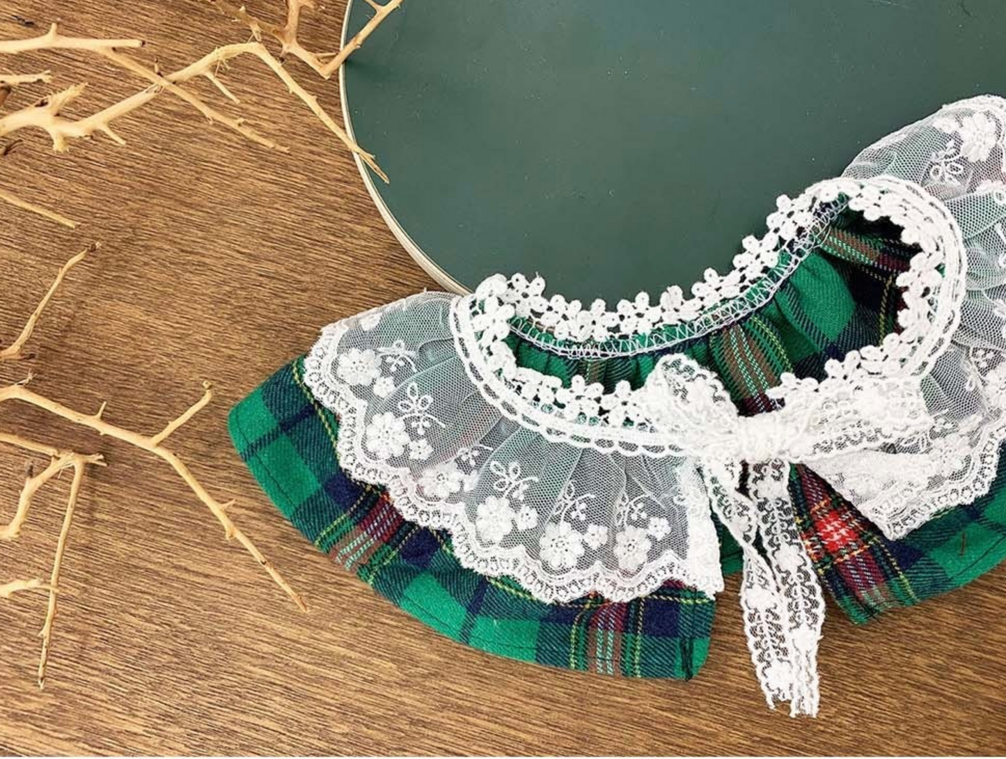 A green plaid dog cape with the same white lace collar and bow detail, arranged flat on a green plate. Rustic wooden background and natural twigs enhance the styling.
