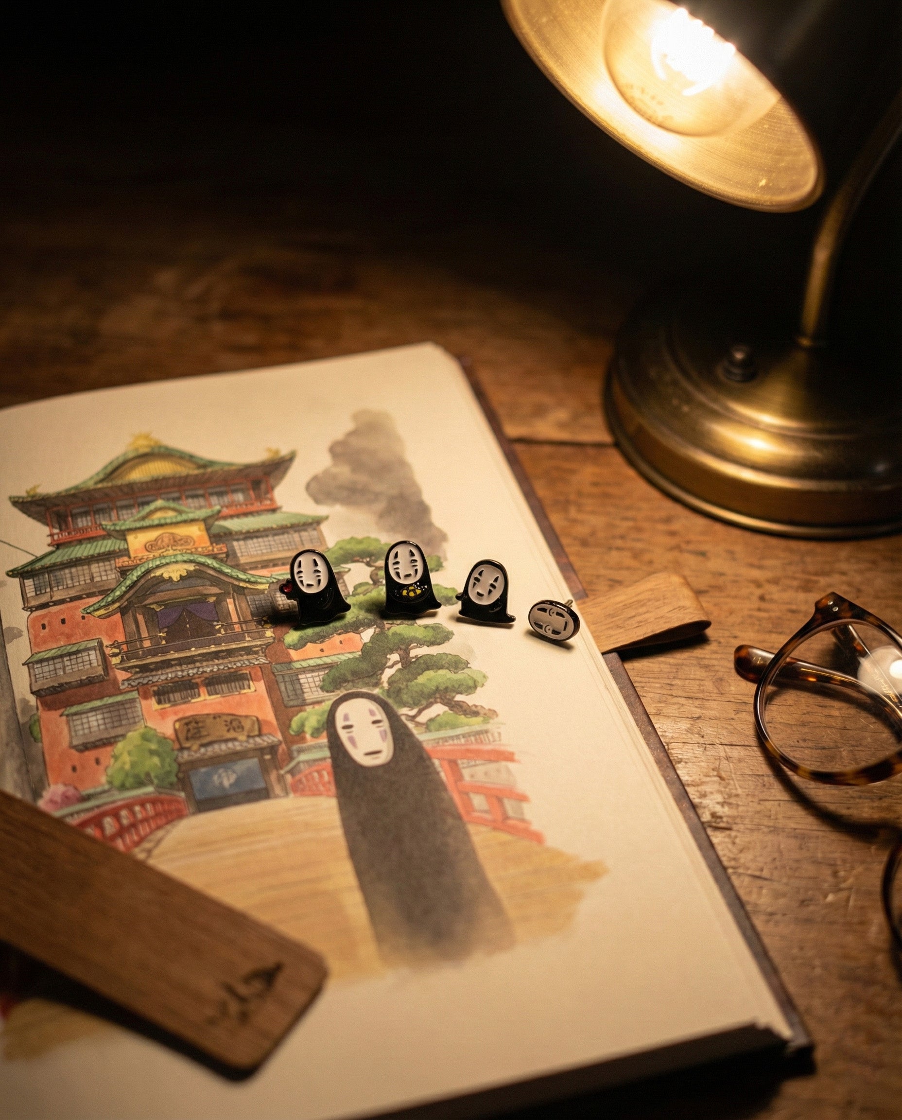  Artistic flat lay of No-Face earrings displayed on illustrated Spirited Away bathhouse postcard with vintage desk lamp and glasses, aesthetic product photography.