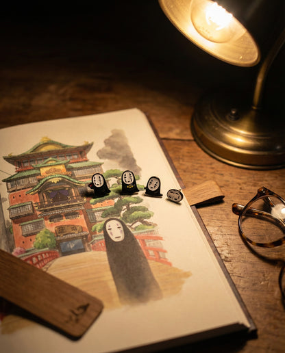  Artistic flat lay of No-Face earrings displayed on illustrated Spirited Away bathhouse postcard with vintage desk lamp and glasses, aesthetic product photography.